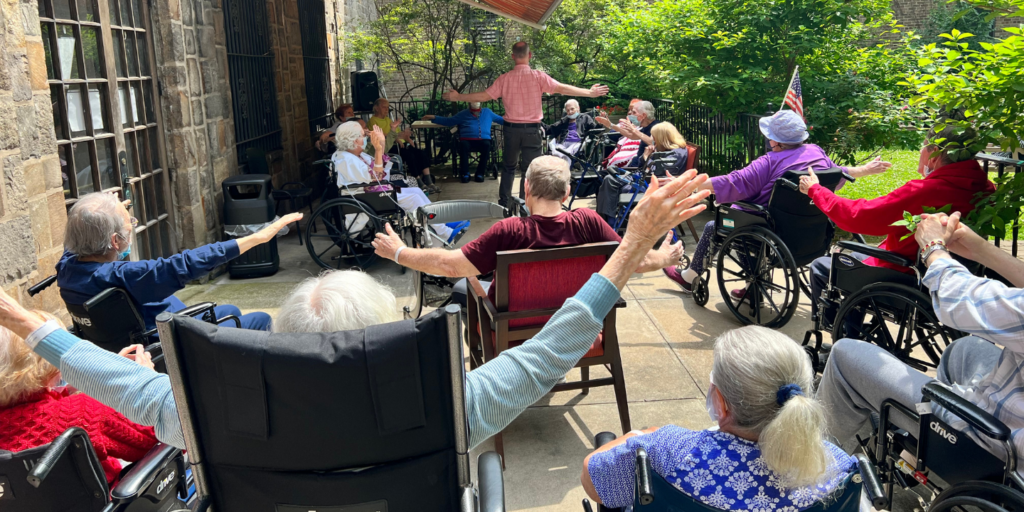 Photo of a group of nursing home residents enjoying a morning stretch routine on the outdoor patio, being led by a Methodist Home team member.