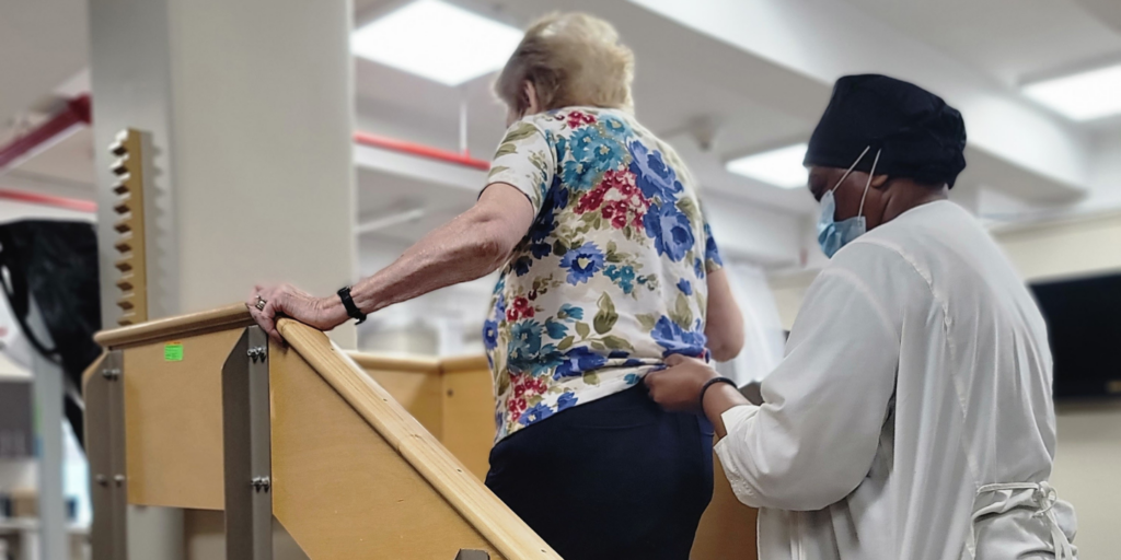 Female short term rehabilitation patient walking up a set of stairs during therapy session, with physical therapist behind her supporting her waist as a guide.