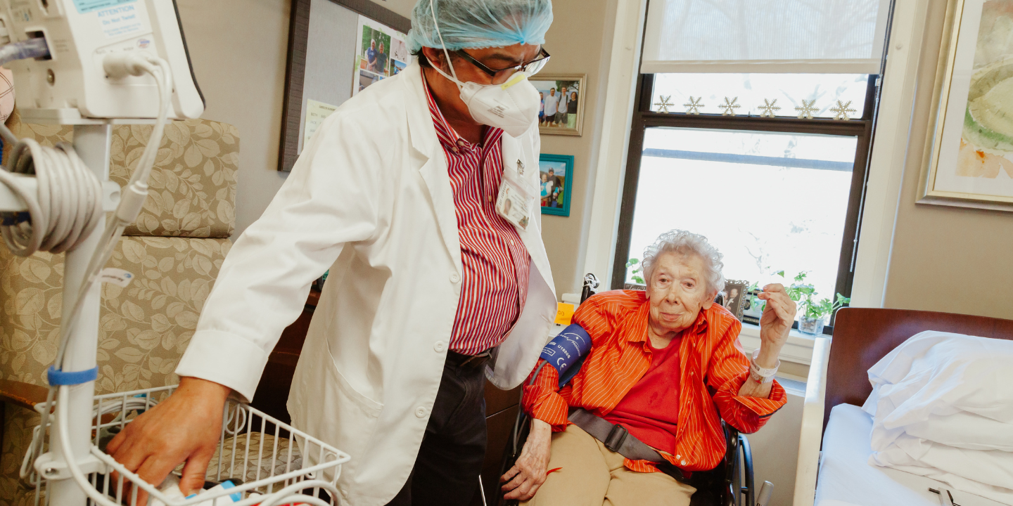 Photo of a female short term rehab patient in her private room getting her blood pressure checked by a physician. The woman looks comfortable and is lightly smiling while looking at the camera.